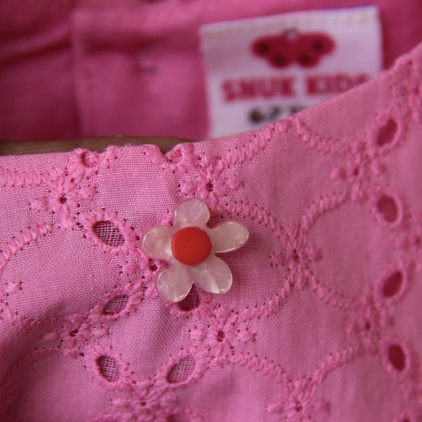 Close-up of pink fabric with a floral button and 'SMUK KIDS' label in the background