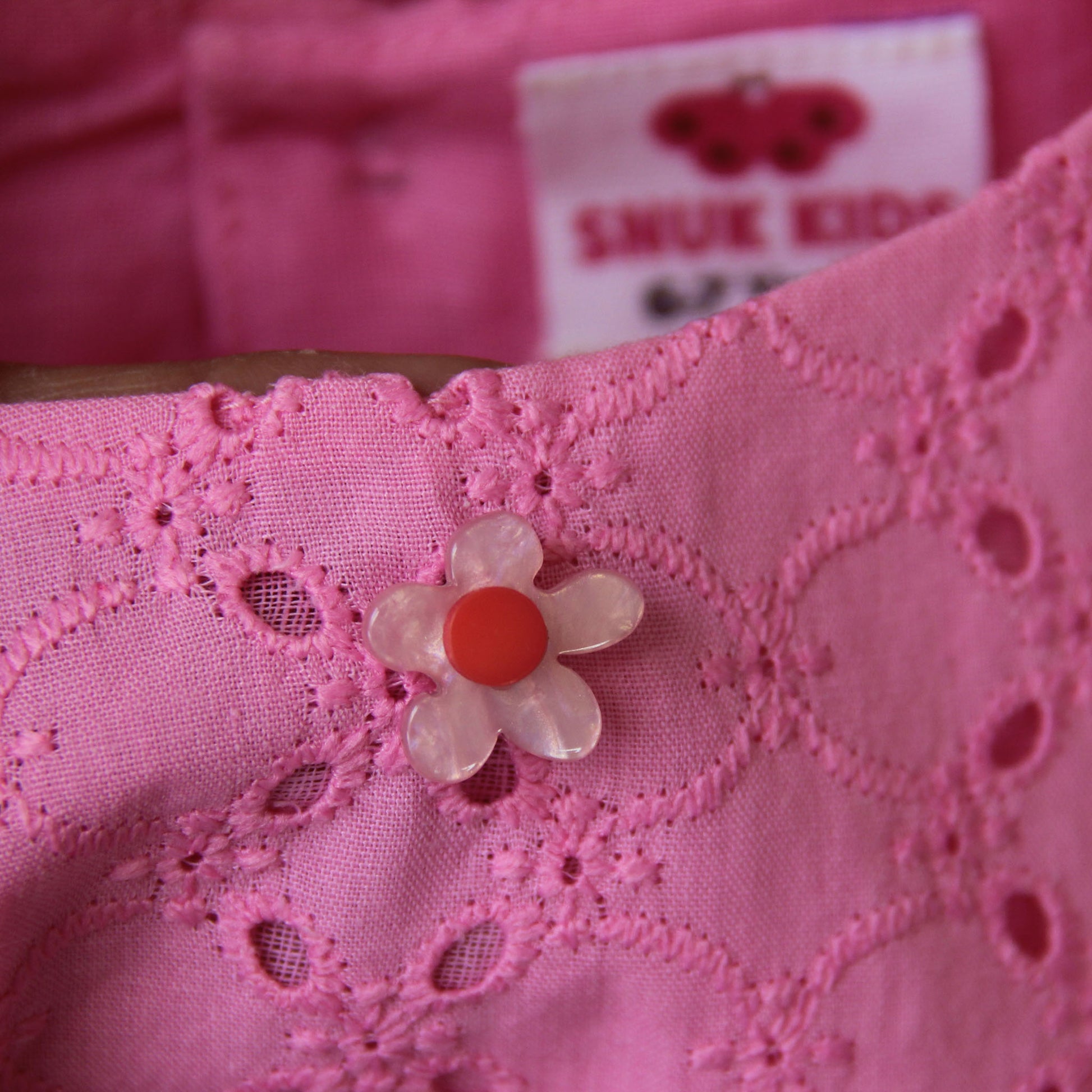 Close-up of pink fabric with a floral button and 'SMUK KIDS' label in the background